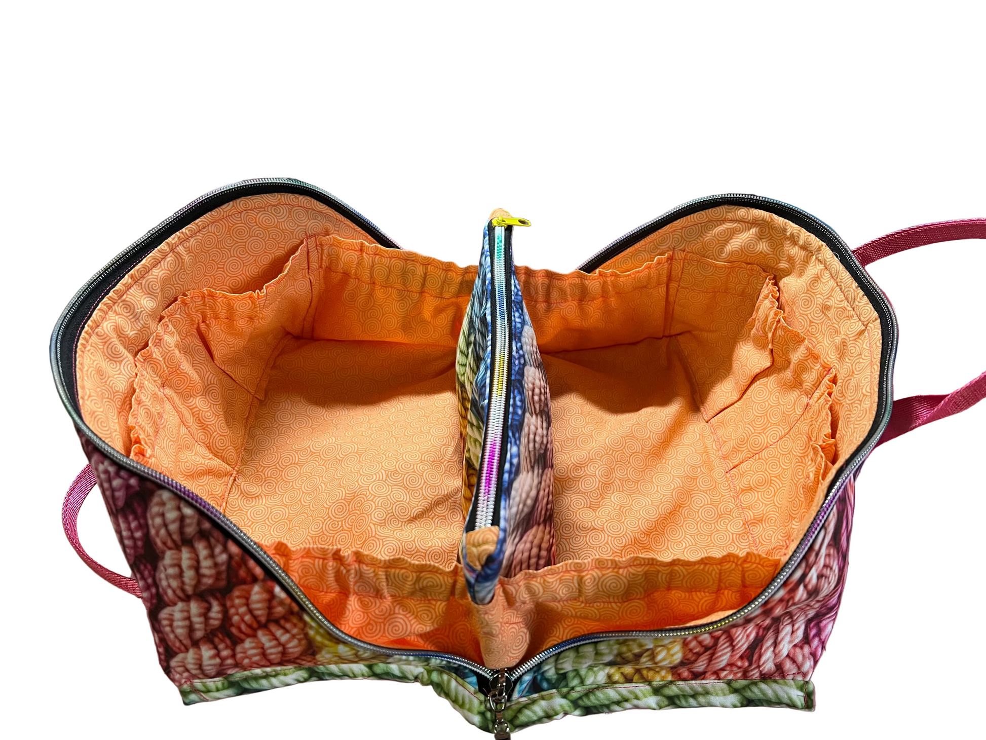 An open zippered fabric organizer bag with a bright orange interior lining and a multicolor patterned exterior, shown on a pink cutting mat in a sewing room setting.