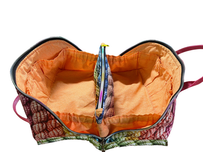 An open zippered fabric organizer bag with a bright orange interior lining and a multicolor patterned exterior, shown on a pink cutting mat in a sewing room setting.